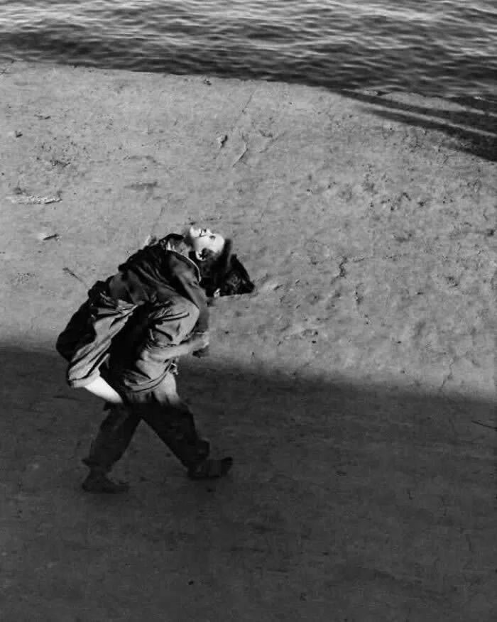 Child carrying another child on their back along a waterfront promenade, with water in the background and long shadows across the pavement, captured from above in a vintage black-and-white photograph.