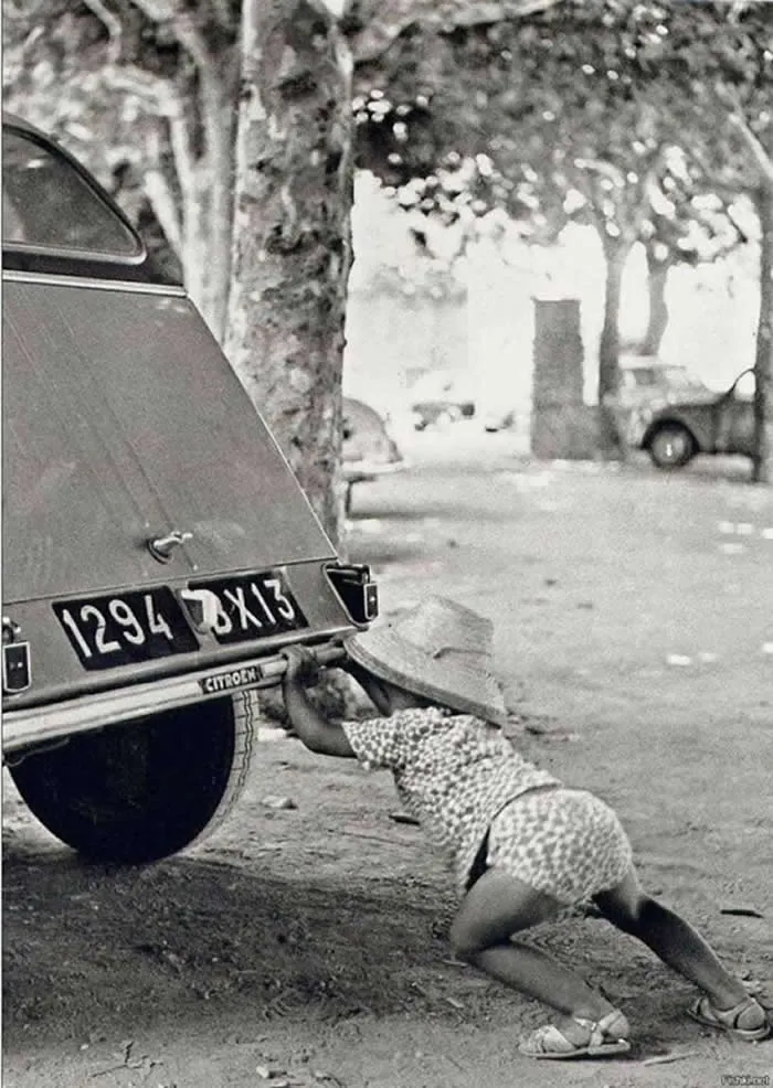 Young child lying on the ground and reaching beneath the rear of a vintage Citro&euml;n car parked on a tree-lined street, captured in a black-and-white photograph.