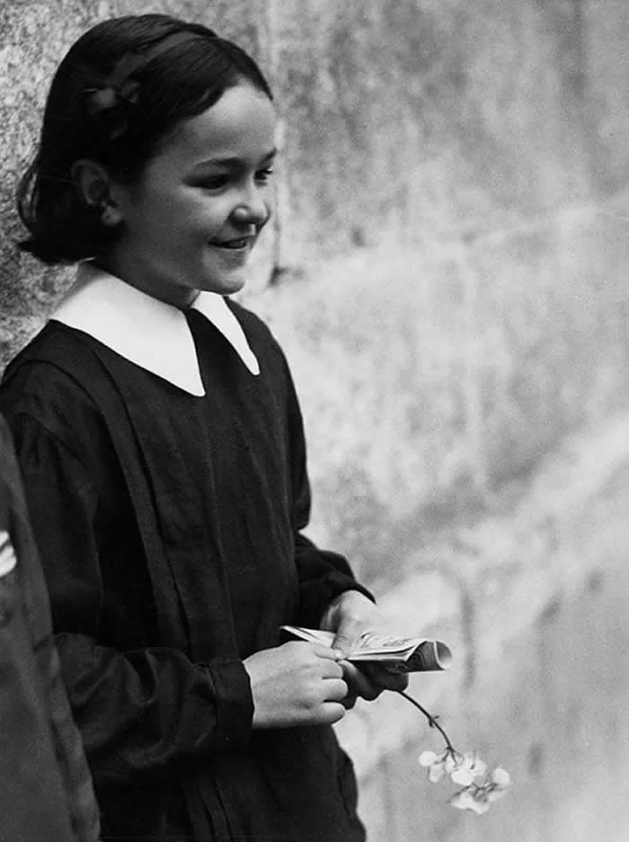 Young schoolgirl in a dark uniform with a white collar, smiling while holding a small book and flowers against a textured wall in a vintage black-and-white portrait.