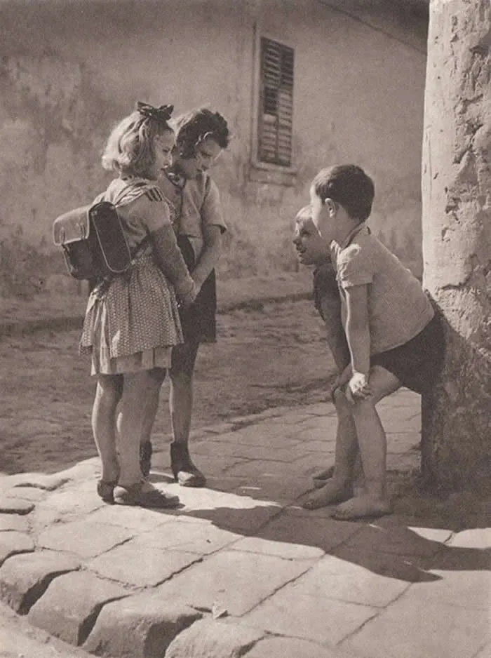 Four children talking together on a sunlit cobblestone street, with two wearing school bags and a rustic building behind them, captured in a vintage monochrome documentary photograph.