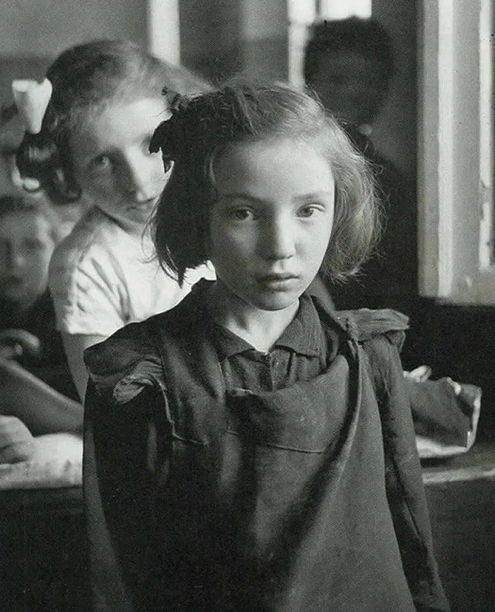Young girl standing in the foreground with two children behind her, photographed indoors with soft natural light in a vintage black-and-white documentary portrait.