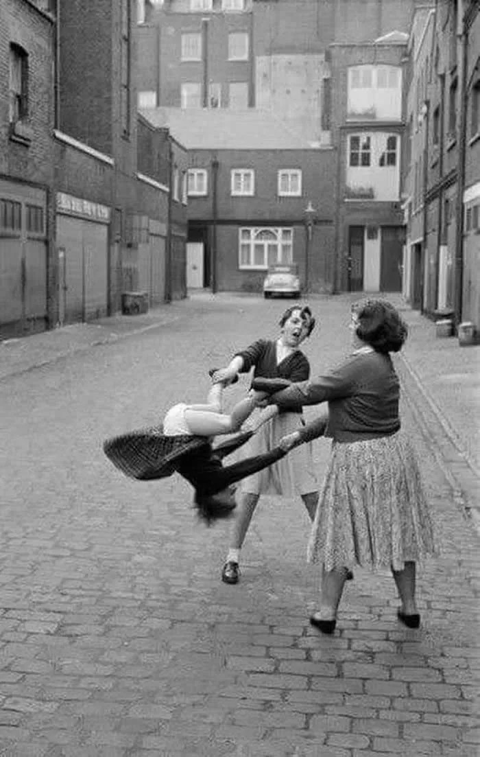 Two children playfully swinging another child through a cobblestone street, surrounded by old brick buildings, captured in a vintage black-and-white photograph.