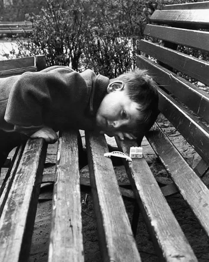 Young child sleeping across a wooden park bench outdoors, with trees and another bench in the background, captured in a candid vintage black-and-white photograph.