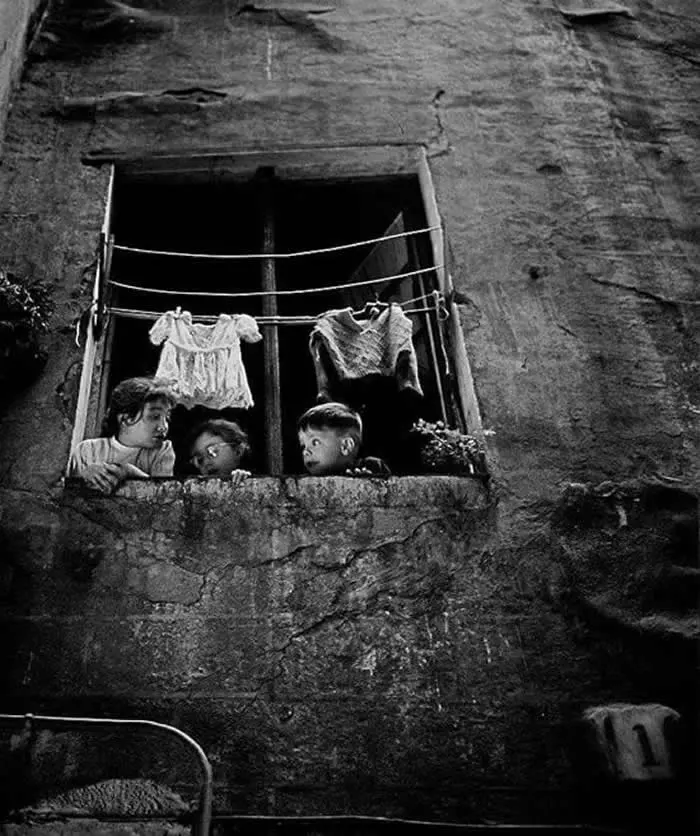 Three children looking out from a weathered window with clothes hanging above them, surrounded by a textured stone wall, captured in a vintage black-and-white documentary photograph.