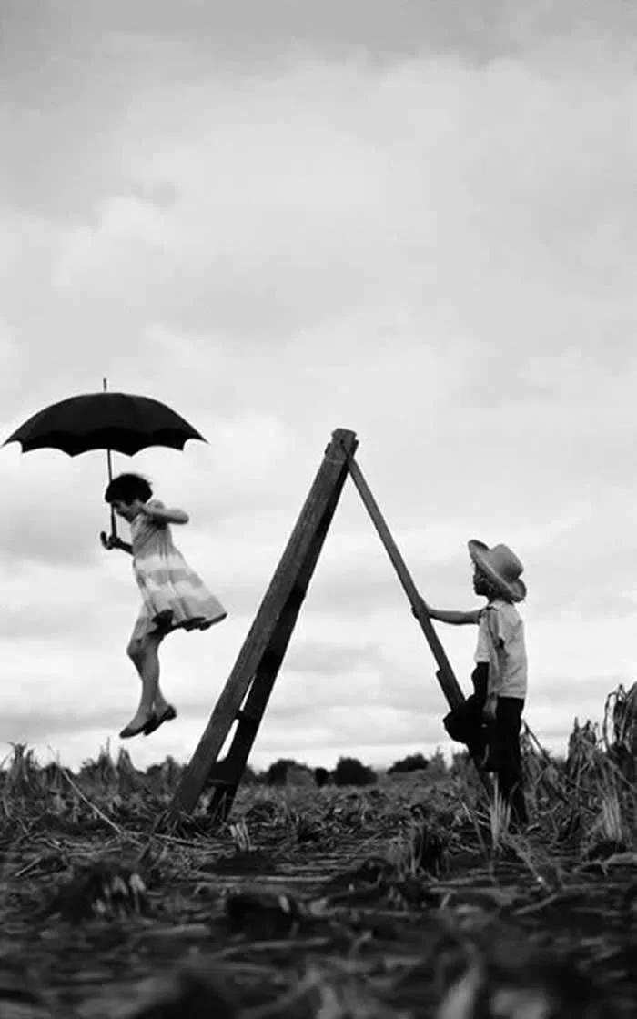 Two children playing on a rustic swing in an open field, with one child holding an umbrella while swinging and the other standing beside the wooden structure, captured in black and white.