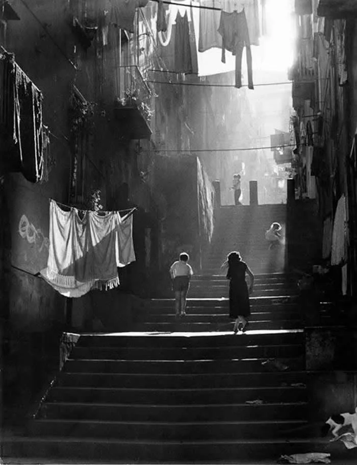 Children walking up a narrow staircase in a dense neighborhood, surrounded by hanging laundry and weathered buildings, with dramatic sunlight illuminating the steps in a vintage black-and-white photograph.