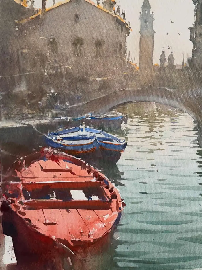 Watercolor painting by Croatian artist Joseph Zbukvic titled &ldquo;Boats,&rdquo; featuring red and blue boats floating along a Venetian canal, with a stone bridge, historic buildings, and a distant bell tower reflected in the water.