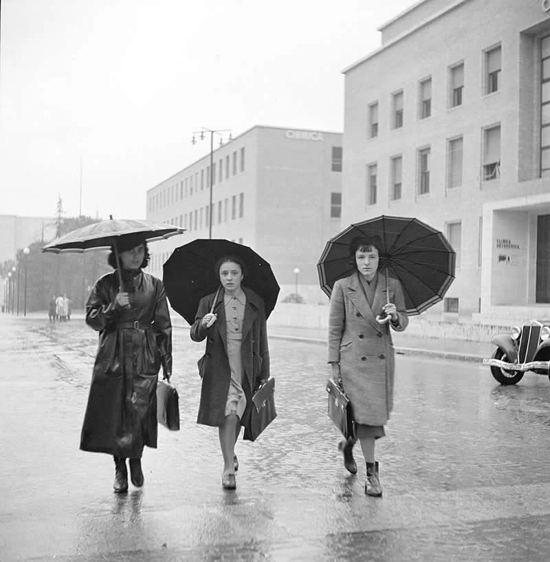 Willem van de Poll: The Master Photographer Who Captured a Changing World 40 Black-and-white photograph of three women carrying umbrellas and handbags while walking along a wet city street in the rain, with tall buildings and a vintage car in the background.