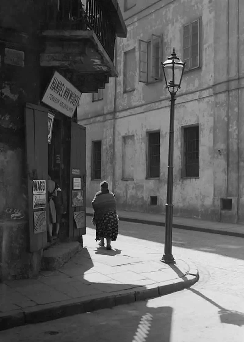 Willem van de Poll: The Master Photographer Who Captured a Changing World 42 Black-and-white photograph of an elderly woman walking past a small shop on a quiet cobblestone street, surrounded by weathered buildings, shuttered windows, and a tall vintage streetlamp casting a long shadow across the sunlit pavement.