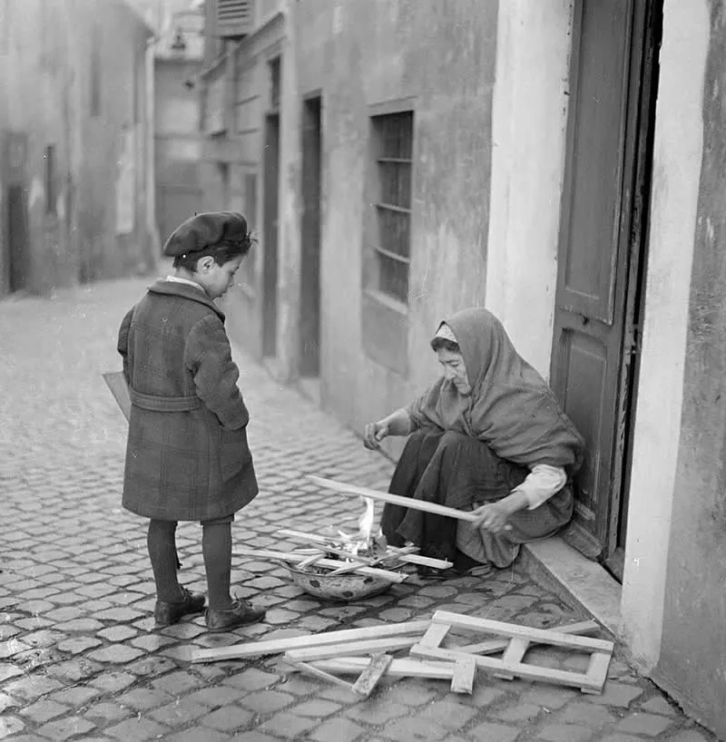 Willem van de Poll: The Master Photographer Who Captured a Changing World 43 Black-and-white photograph of a young boy in a long coat and cap watching an elderly woman seated in a doorway, arranging wooden sticks over a small fire on a cobblestone street.