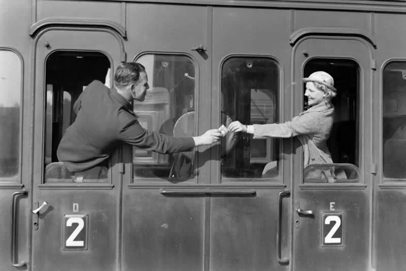 Willem van de Poll: The Master Photographer Who Captured a Changing World 44 Black-and-white photograph of a man leaning out of a train window to exchange a small item with a woman leaning from another window, with numbered second-class compartments and rectangular windows forming a balanced composition.