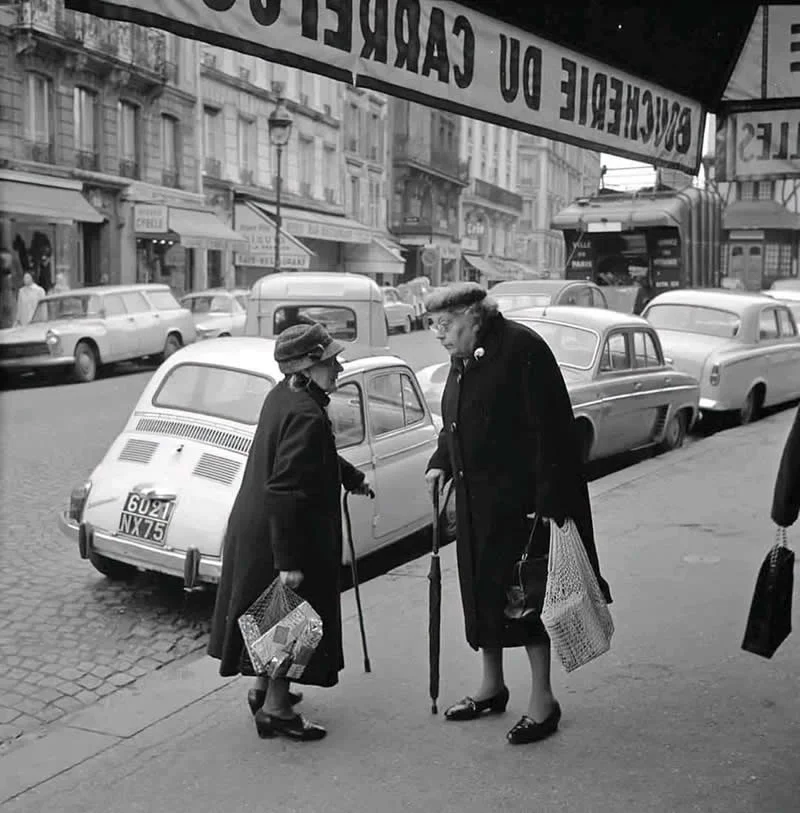 Willem van de Poll: The Master Photographer Who Captured a Changing World 45 Black-and-white photograph of two elderly women in dark coats and hats chatting on a city sidewalk, carrying shopping bags and umbrellas, with vintage cars, storefronts, and a large shop awning in the background.