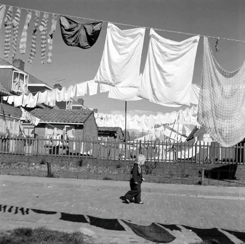 Willem van de Poll: The Master Photographer Who Captured a Changing World 47 Black-and-white photograph of a young child walking along a paved street beneath clotheslines filled with white sheets and garments, with modest houses, tiled rooftops, a wooden fence, and long shadows in the foreground.