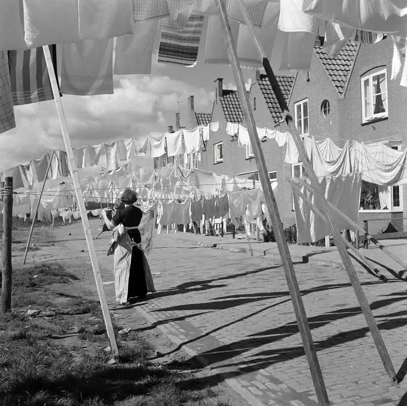 Willem van de Poll: The Master Photographer Who Captured a Changing World 48 Black-and-white photograph of a woman carrying laundry beneath numerous clotheslines filled with hanging sheets and garments in a residential street, with brick houses, tiled roofs, and long shadows stretching across the pavement.