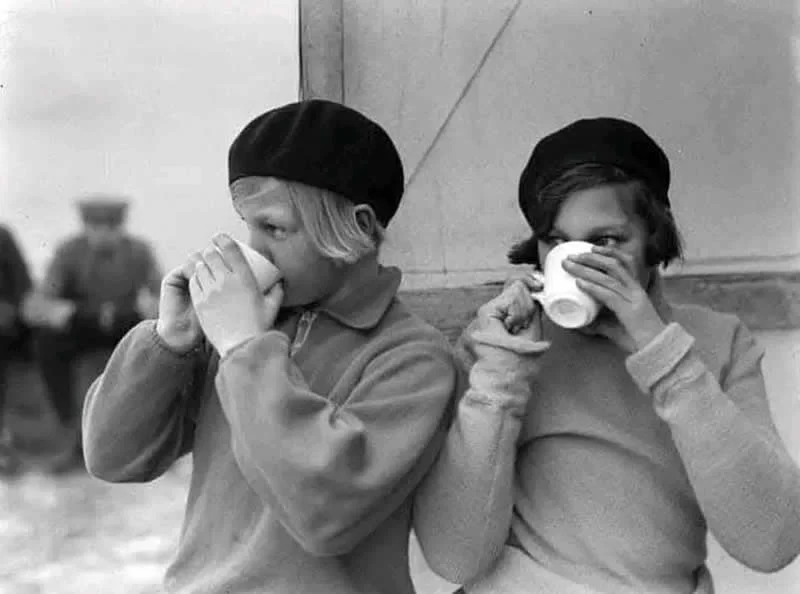 Willem van de Poll: The Master Photographer Who Captured a Changing World 49 Black-and-white photograph of two young women wearing dark berets and coats, drinking from white cups while sitting beside a wall, with a blurred figure visible in the background.