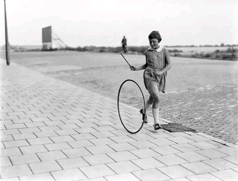 Willem van de Poll: The Master Photographer Who Captured a Changing World 50 Black-and-white photograph of a young girl in a sleeveless dress and knee-high socks rolling a large hoop with a stick across a paved waterfront promenade, with open water and a distant figure in the background.