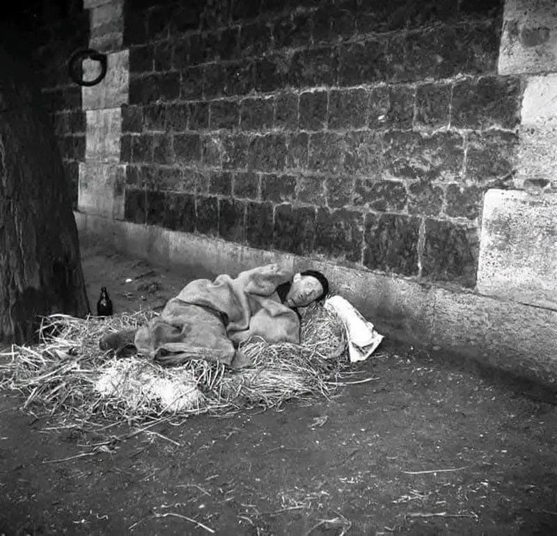 Willem van de Poll: The Master Photographer Who Captured a Changing World 51 Black-and-white photograph of a person sleeping on straw beneath a tree, wrapped in a blanket beside a weathered stone wall, with a small bottle nearby.
