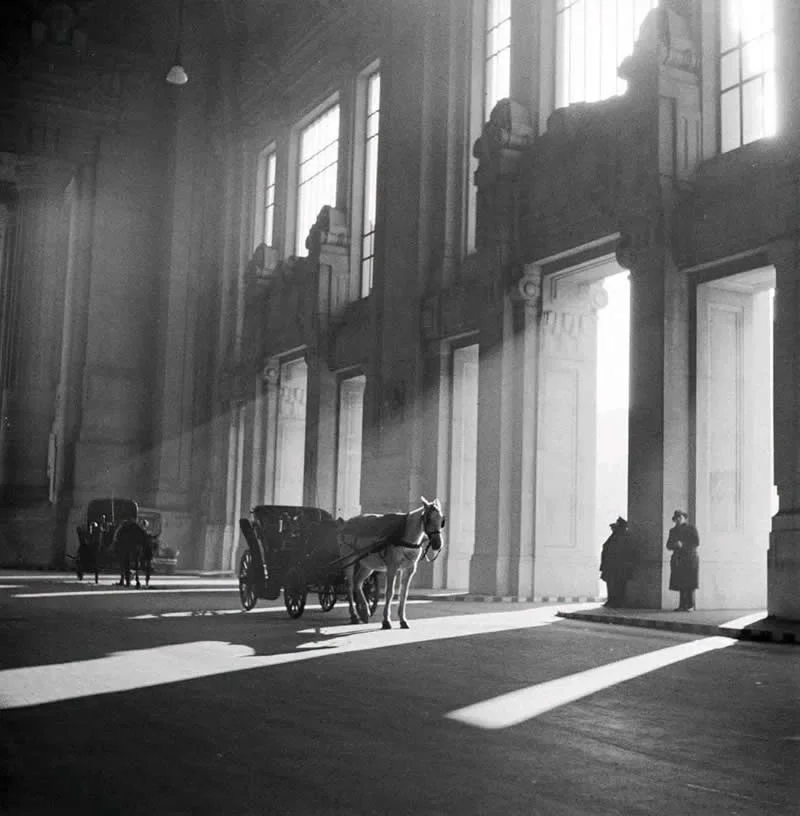 Willem van de Poll: The Master Photographer Who Captured a Changing World 53 Black-and-white photograph of horse-drawn carriages and a few people inside a grand hall with towering windows and monumental columns, where long beams of sunlight cast dramatic shadows across the floor.