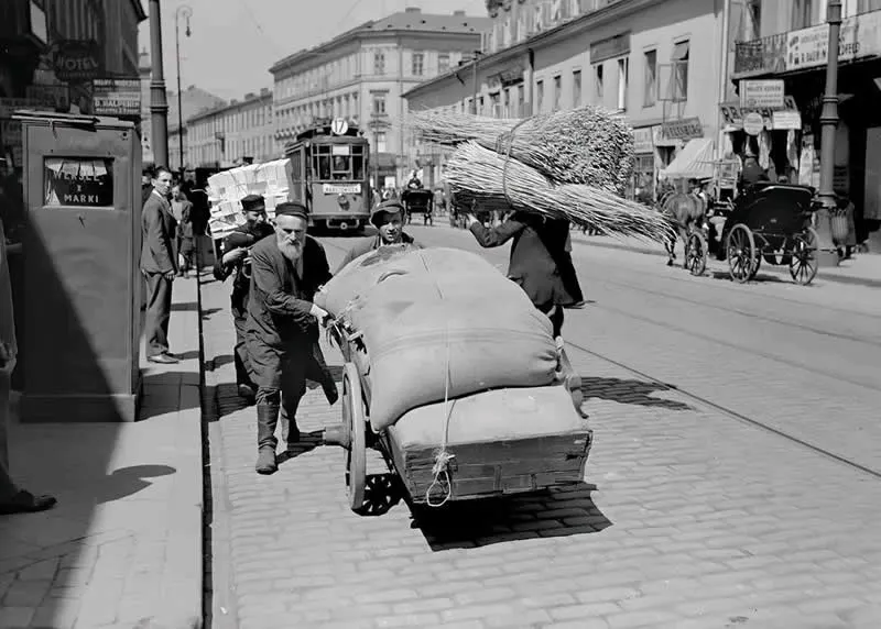 Willem van de Poll: The Master Photographer Who Captured a Changing World 54 Black-and-white photograph of men pushing a heavily loaded handcart along a cobblestone street, with a tram, horse-drawn carriages, pedestrians, and historic European buildings filling the busy urban background.