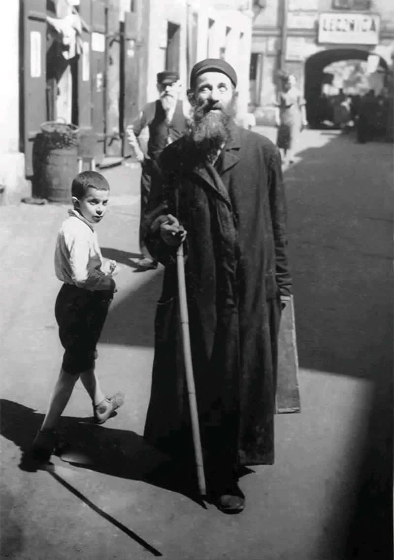 Willem van de Poll: The Master Photographer Who Captured a Changing World 55 Black-and-white photograph of an elderly bearded man wearing a cap and long coat, holding a walking stick on a European street, while a young boy walks past behind him and pedestrians appear in the background.