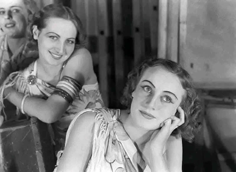 Black-and-white photograph of two women wearing patterned dresses, with the woman in front resting her head on her hand and gazing toward the camera while another woman smiles behind her.