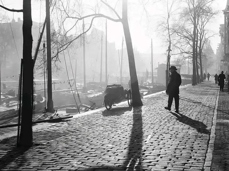 Willem van de Poll: The Master Photographer Who Captured a Changing World 57 Black-and-white photograph of a man walking along a cobblestone street beneath leafless trees, with horse-drawn carts, distant pedestrians, and historic buildings fading into mist. Long tree shadows stretch dramatically across the sunlit pavement.