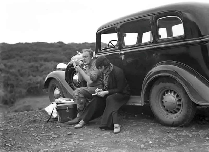Willem van de Poll: The Master Photographer Who Captured a Changing World 58 Black-and-white photograph of two men sitting beside a vintage car on a rural roadside. One man leans toward the vehicle while the other looks down at a paper, with a low, wooded hillside stretching across the background.