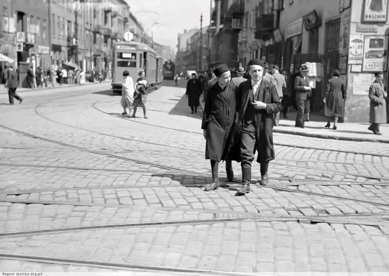 Willem van de Poll: The Master Photographer Who Captured a Changing World 60 Black-and-white photograph of two men wearing long coats and caps walking arm in arm across tram tracks on a cobblestone street, with a tram, pedestrians, and historic European buildings in the background.