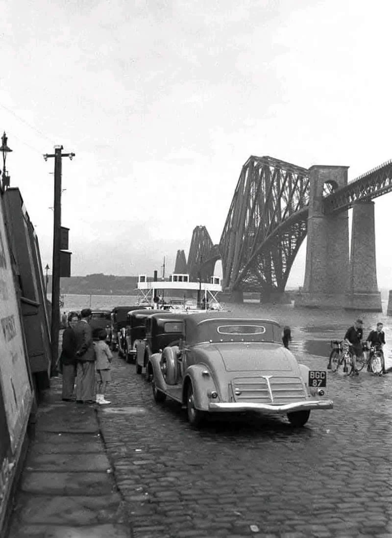 Willem van de Poll: The Master Photographer Who Captured a Changing World 61 Black-and-white photograph of vintage cars parked along a cobblestone waterfront beneath a massive steel railway bridge, with pedestrians gathered beside the vehicles and two cyclists riding near the water.