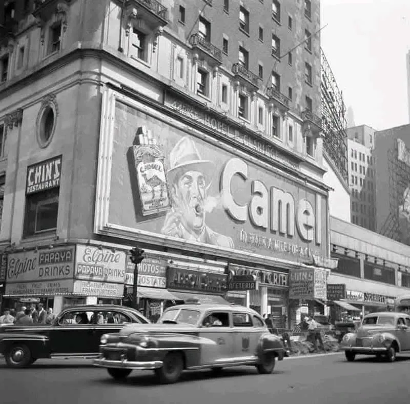 Willem van de Poll: The Master Photographer Who Captured a Changing World 62 Black-and-white photograph of a busy urban street lined with tall buildings and storefronts, featuring a large Camel cigarette billboard above the shops as vintage cars drive past pedestrians along the road.