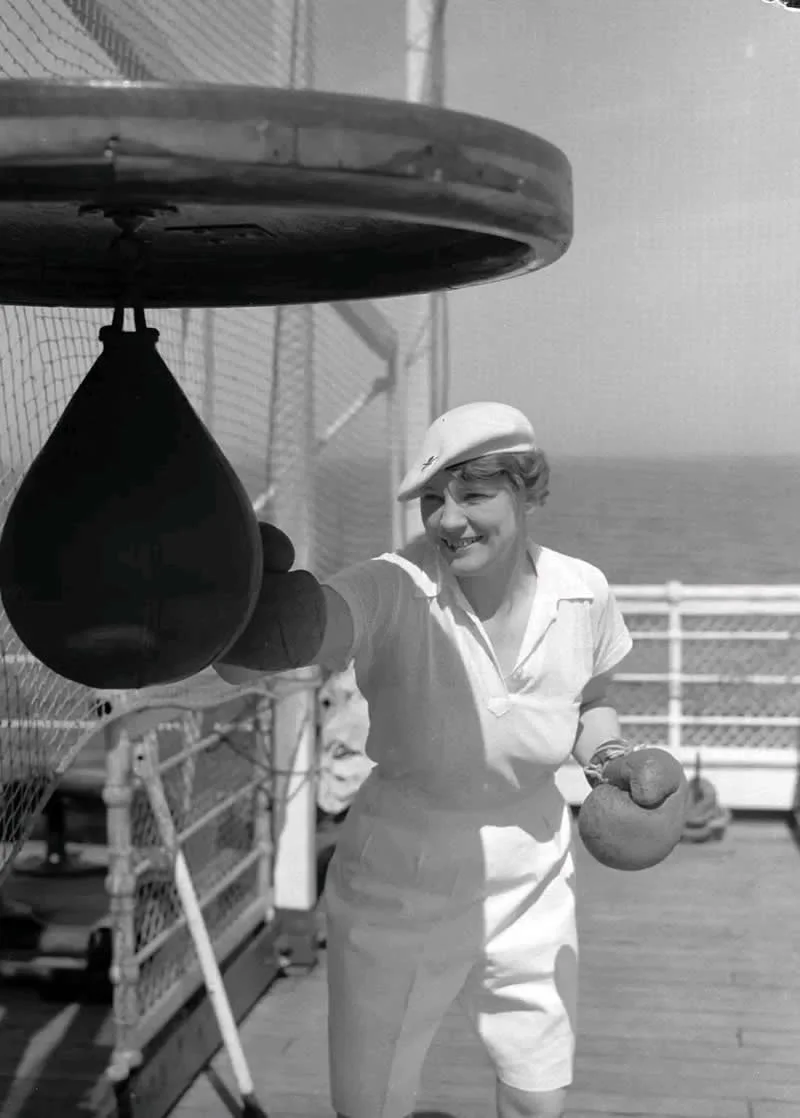 Willem van de Poll: The Master Photographer Who Captured a Changing World 63 Black-and-white photograph of a smiling woman wearing a white sports outfit, cap, and boxing gloves as she punches a hanging speed bag on a ship’s deck, with metal railings and open water behind her.