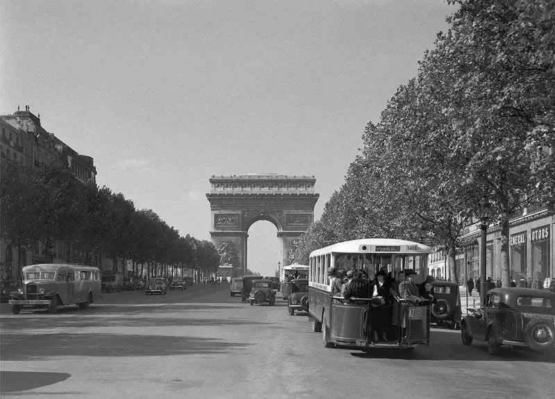 Willem van de Poll: The Master Photographer Who Captured a Changing World 64 Black-and-white photograph of a busy Parisian boulevard leading toward the Arc de Triomphe, with vintage buses, classic cars, and pedestrians beneath rows of leafy trees lining the broad avenue.