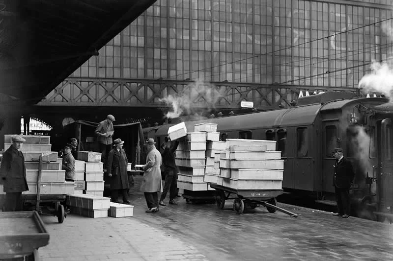 Willem van de Poll: The Master Photographer Who Captured a Changing World 65 Black-and-white photograph of railway workers handling stacks of boxes on luggage carts beside a passenger train at a large station, with steam rising around the train and a metal-and-glass station structure in the background.