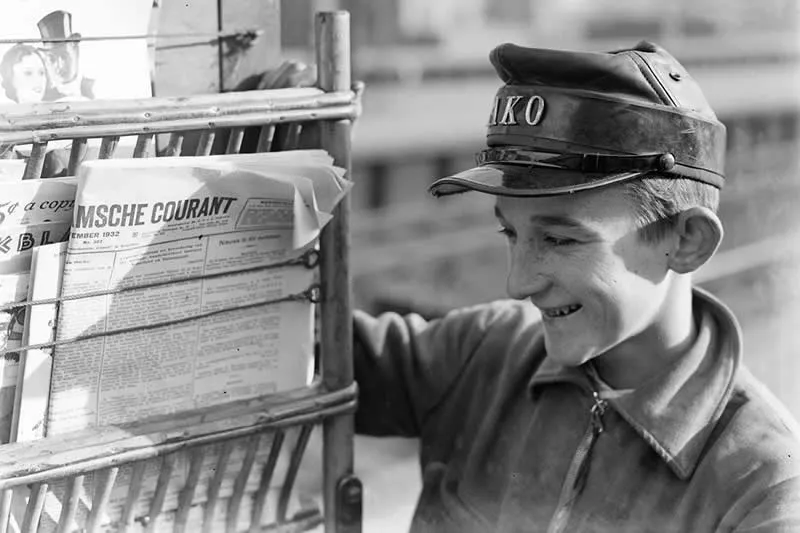 Willem van de Poll: The Master Photographer Who Captured a Changing World 66 Black-and-white photograph of a smiling young newspaper seller wearing a peaked cap and jacket, standing beside a metal newsstand filled with folded newspapers, including a prominently displayed French newspaper.