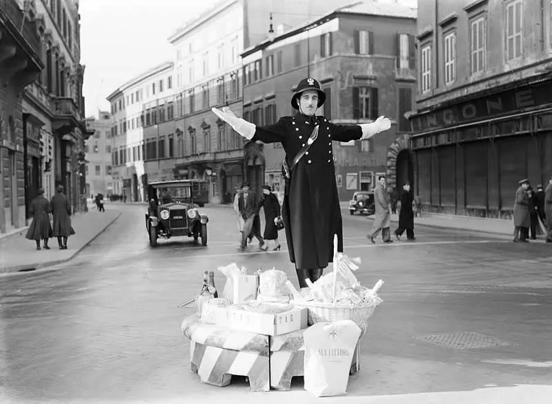 Willem van de Poll: The Master Photographer Who Captured a Changing World 67 Black-and-white photograph of a uniformed man wearing a peaked hat and long coat, standing with arms outstretched on a street display piled with goods, surrounded by historic European buildings, vintage cars, and pedestrians.