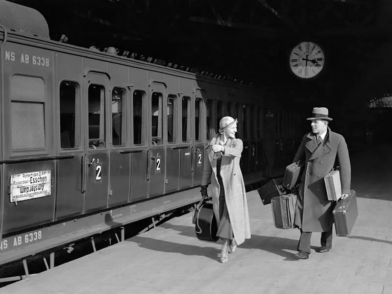 Willem van de Poll: The Master Photographer Who Captured a Changing World 69 Black-and-white photograph of a well-dressed man and woman carrying luggage beside a vintage passenger train at a railway station, with a large clock mounted above the platform and sunlight casting dramatic shadows across the scene.