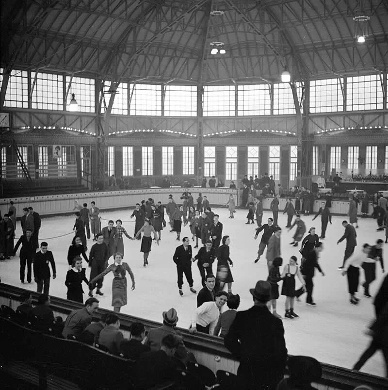 Willem van de Poll: The Master Photographer Who Captured a Changing World 71 Black-and-white photograph of men and women ice-skating at a crowded indoor rink beneath a high steel-truss roof with large windows, while spectators watch from wooden benches along the rink’s edge.