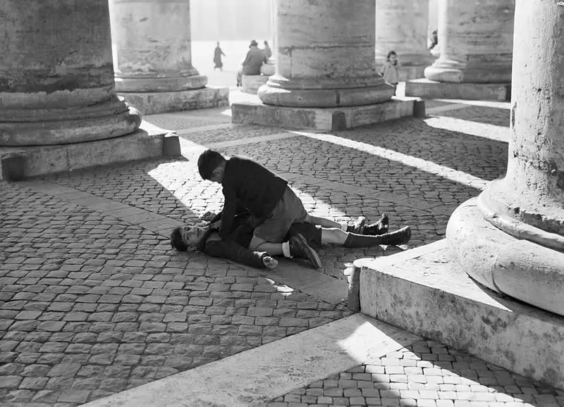 Willem van de Poll: The Master Photographer Who Captured a Changing World 72 Black-and-white photograph of two people lying on cobblestones beneath massive stone columns, with one person leaning over the other. Strong sunlight casts long geometric shadows across the pavement, while distant pedestrians appear between the columns.