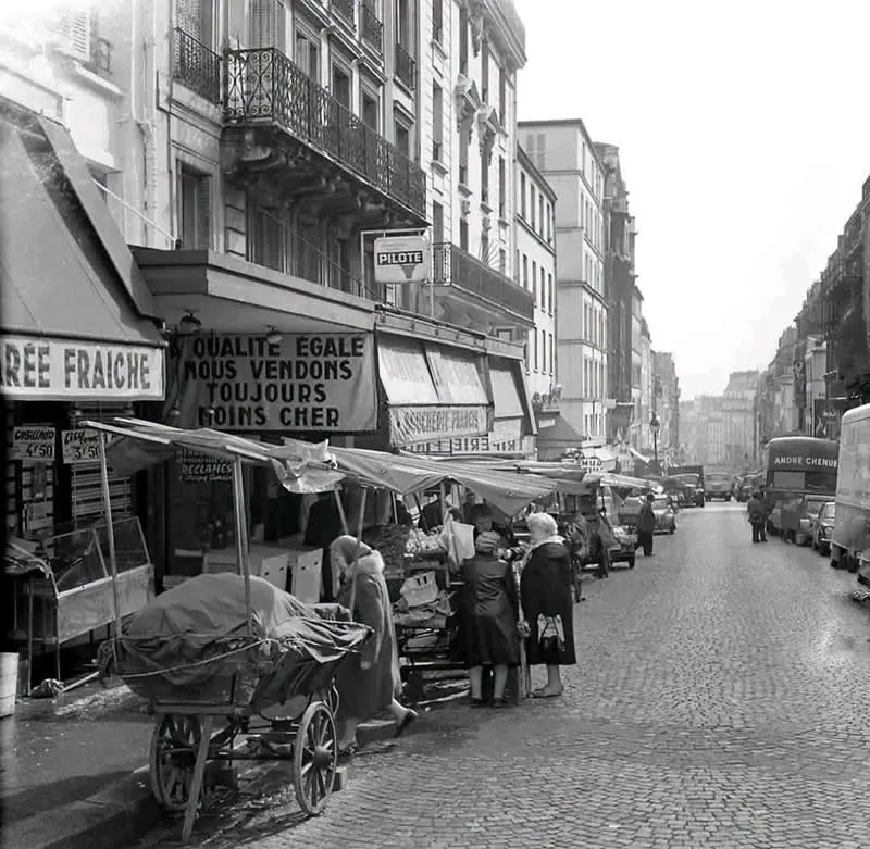 Willem van de Poll: The Master Photographer Who Captured a Changing World 74 Black-and-white photograph of shoppers browsing outdoor market stalls beneath awnings along a cobblestone street lined with historic European buildings, vendor carts, parked vintage vehicles, and pedestrians stretching into the distance.